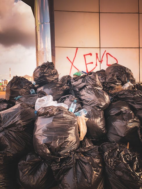 A narrow urban alleyway filled with a large collection of mixed waste materials and rubbish. In the foreground, a large, worn, grey refuse bag or sack, possibly made of heavy-duty plastic or woven fabric, is positioned on the uneven, dark asphalt ground, showing signs of use and slight dirt. Behind it, there are discarded items, including a small black trash bin lying on its side, and various pieces of refuse scattered around. Further back, a dense pile of cardboard boxes, paper, and plastic waste is stacked against a weathered brick wall with some graffiti on it. To the sides, the alley is bordered by aged buildings with a mix of brick, metal, and plaster surfaces, some with visible graffiti or paint marks. Bare tree branches extend above, suggesting the scene is set in late autumn or winter, with muted natural lighting casting minimal shadows. The environment appears to be a neglected alley used for waste disposal, illustrating an instance of private or independent rubbish collection in an urban setting, as handled by local waste removal services like House Clearance Knightsbridge, prior to scheduled clearance or disposal.