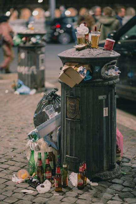 A black, cylindrical metal public trash bin situated on a cobblestone street, with various litter occurrences around its base and on top. The bin features a textured, slightly weathered surface, with a small rectangular slot near the top for trash disposal and a more extensive opening on the side, partially obscured. On the top, there are several discarded beverage cups, some with plastic lids and straws, a crumpled paper napkin, and a small, offset cardboard box. Surrounding the bin, the ground is strewn with numerous empty glass bottles, some upright and others knocked over, along with crumpled paper, plastic wrappers, and other miscellaneous trash. In the background, a blurred scene of people walking and standing, with parked cars and additional rubbish, can be seen, indicating a busy urban environment. The overall scene captures the impact of littering in a public space, highlighting the importance of proper waste disposal, and situating the image within the context of waste management and rubbish removal services like those offered by House Clearance Knightsbridge.