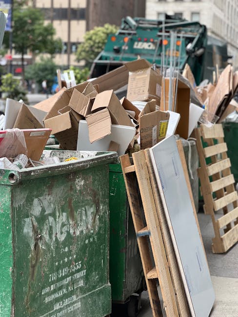 A large green waste collection bin filled with a variety of discarded cardboard boxes, some flattened and others partially intact, alongside wooden pallets leaning against the side of the bin. The cardboard is in different shades of brown, with some boxes showing printed labels or tape remnants. In the background, a skip loader vehicle with a blue-green body and controlling arm is visible, situated on a paved urban street with multi-story buildings and green trees. The scene is outdoors during daytime, under overcast lighting, illustrating an area prepared for rubbish removal or private waste collection involving cardboard and wooden debris, consistent with alternative waste handling services like those offered by House Clearance Knightsbridge.