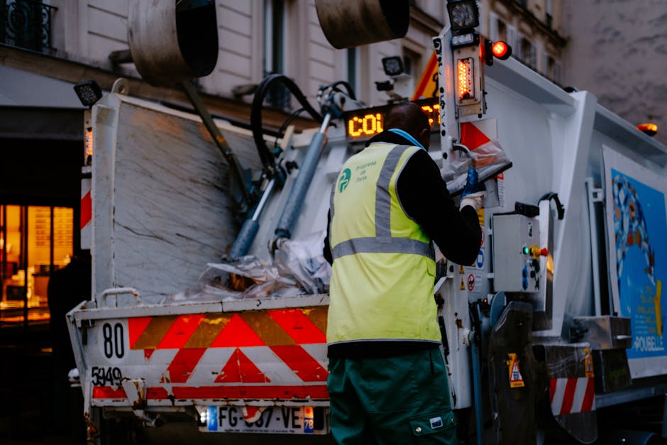 A waste collection worker wearing a high-visibility yellow vest and dark clothing is operating an underground waste compaction vehicle with a digital display showing 'CO1' on the back. The vehicle's large rear hopper is open and filled with mixed rubbish, including paper, plastic, and other refuse materials. The vehicle is parked on a city street, with a building visible in the background, illuminated by warm interior lighting. The scene is captured during dusk or early evening, with artificial lighting highlighting the reflective markings on the waste truck. This setup exemplifies a form of private or alternative waste handling, in contrast to standard bin collection, often associated with independent waste disposal services like those provided by House Clearance Knightsbridge, which manage rubbish removal in densely populated urban areas, such as Knightsbridge, ensuring proper disposal and clearance of waste debris.