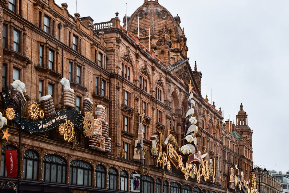 A detailed exterior view of a historic, ornate sandstone building in Knightsbridge, featuring large arched windows and classical architectural detailing. The building is decorated with festive holiday displays, including large, striped candy cane-shaped ornaments, golden star figures, and illuminated decorative elements, some of which resemble oversized confectionery items. The decorations span the building's facade and are mounted on black metal frames attached to the structure. The scene is captured during daytime with an overcast sky, contributing soft, diffuse lighting. The surroundings include a paved street or pedestrian area in the foreground, indicating an urban setting. The elaborate decorations, combined with the classical architecture, reflect a seasonal adornment likely aimed at attracting visitors and shoppers. The image pertains to local property or commercial premises in need of routine rubbish removal, potentially stored or managed by House Clearance Knightsbridge, supporting alternative waste disposal methods outside public authority collection, especially during peak festive periods.