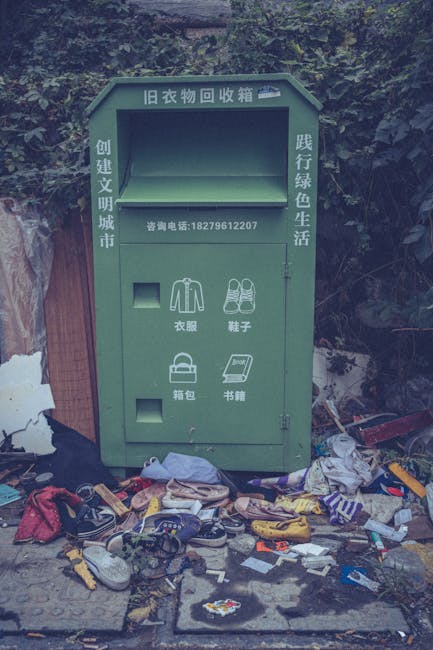 A green, old-fashioned clothing recycling bin with multiple compartments and Chinese characters on its surface, positioned outdoors against a backdrop of dense shrubbery and overgrown plants. The bin has a rectangular shape with a sloped top, and features separate slots labeled for different types of textiles, including garments, shoes, bags, and books, with white icons illustrating each category. In front of the bin, there is a pile of discarded clothing, shoes, and miscellaneous waste items, such as fabric, plastic bags, and small debris, scattered across the ground. The surrounding environment appears untidy, reflecting a lack of regular collection or maintenance, with the clutter extending to the pavement area around the bin. The image is captured during daylight with natural lighting, emphasizing the contrast between the organized recycling structure and the unmanaged waste scattered nearby, illustrating an example of independent waste handling by residents or local disposal services like House Clearance Knightsbridge.