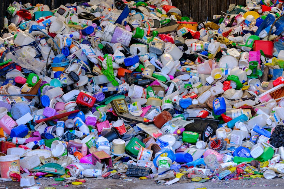 A large outdoor pile of mixed household rubbish consisting of plastic containers, bottles, and caps in various colours including white, blue, green, pink, and red. The debris is scattered across an uneven surface, with some items stacked and overlapping, creating a chaotic scene. Among the waste are plastic tubs, drinking cups, and small packaging remnants, some with labels visible but unreadable. The background features a weathered wooden fence, hinting at an outdoor environment such as a driveway or waste collection area. The overall scene underscores the importance of proper rubbish removal and recycling, reflecting services provided by companies like House Clearance Knightsbridge for alternative waste handling and on-site clearance. Bright natural lighting reveals the textures of the plastic materials, highlighting the volume and variety of household waste typically addressed during professional rubbish collection and disposal tasks.