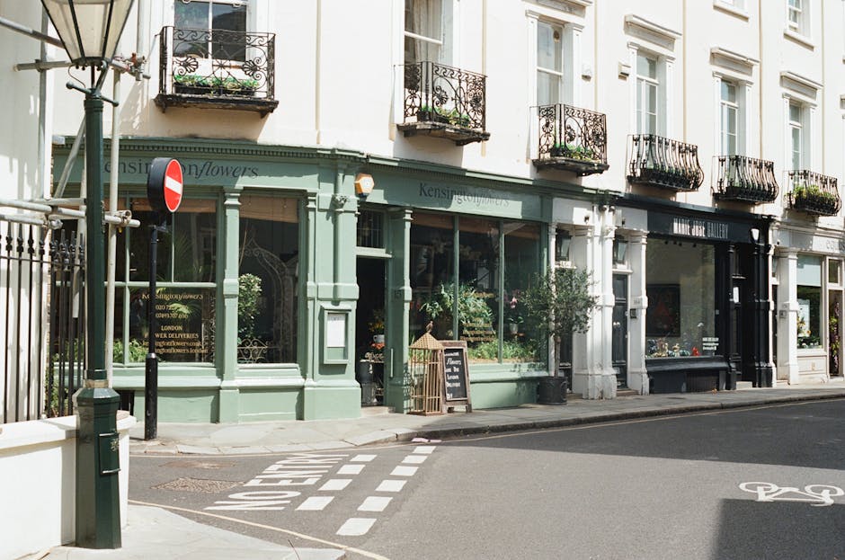 A row of three-story buildings with ground-floor retail spaces and residential flats above, situated along a quiet city street. The storefronts include large glass display windows revealing interior displays of plants and flower arrangements. The first shop, painted in a soft mint green, features a small outdoor chalkboard sign on the pavement, and potted plants placed near the entrance, suggesting it is a flower shop. The other storefronts are painted in white and black, with decorative iron balcony railings on the upper floors, some with small potted plants on the balconies. A street lamp stands on the corner, and a metal pole with a no-entry road sign is visible near the sidewalk. The street has a painted bicycle lane and pedestrian crosswalk markings. The scene is well-lit with natural daylight, and the overall environment suggests a European urban setting with a blend of traditional and modern architectural elements, consistent with typical independent retail premises that might be involved in private waste collection or external rubbish removal services like those offered by House Clearance Knightsbridge.