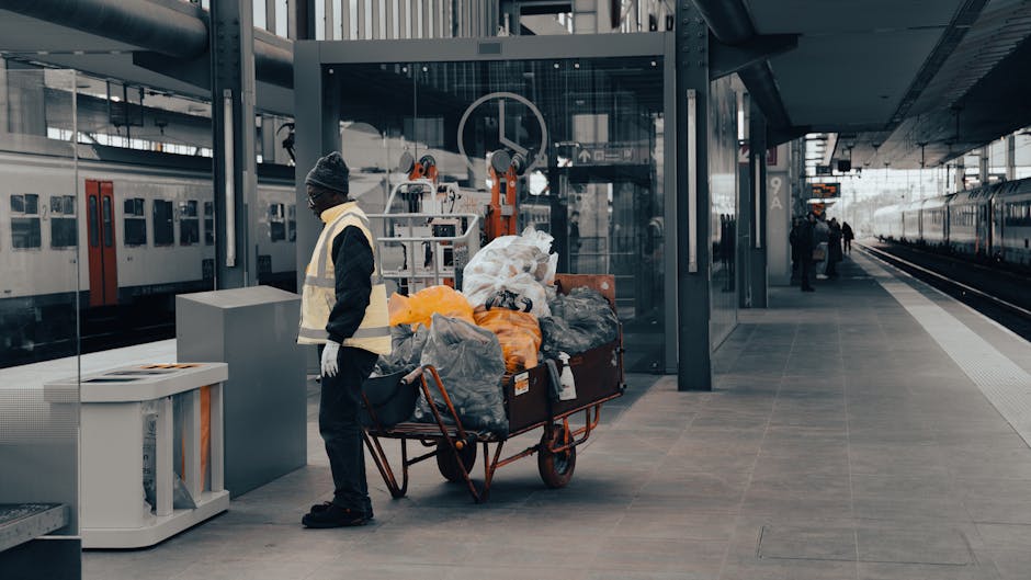 A worker wearing a dark jacket, grey beanie, and reflective safety vest stands next to a large red trolley cart filled with various bags and items of rubbish, including black, grey, and orange plastic garbage bags, along with some loose garbage. The trolley is positioned on a paved platform at an urban train station, with modern glass and metal structures surrounding the area. In the background, trains are visible on adjacent tracks, with a few other commuters walking on the platform or waiting nearby. The environment is well-lit with natural daylight, highlighting the textures of the bags and the smooth, polished surface of the platform. This scene illustrates a typical setting where rubbish or waste is temporarily accumulated before disposal or transfer, aligning with private waste handling and rubbish removal services such as those offered by House Clearance Knightsbridge, within the context of alternative waste management logistics at a busy railway station.