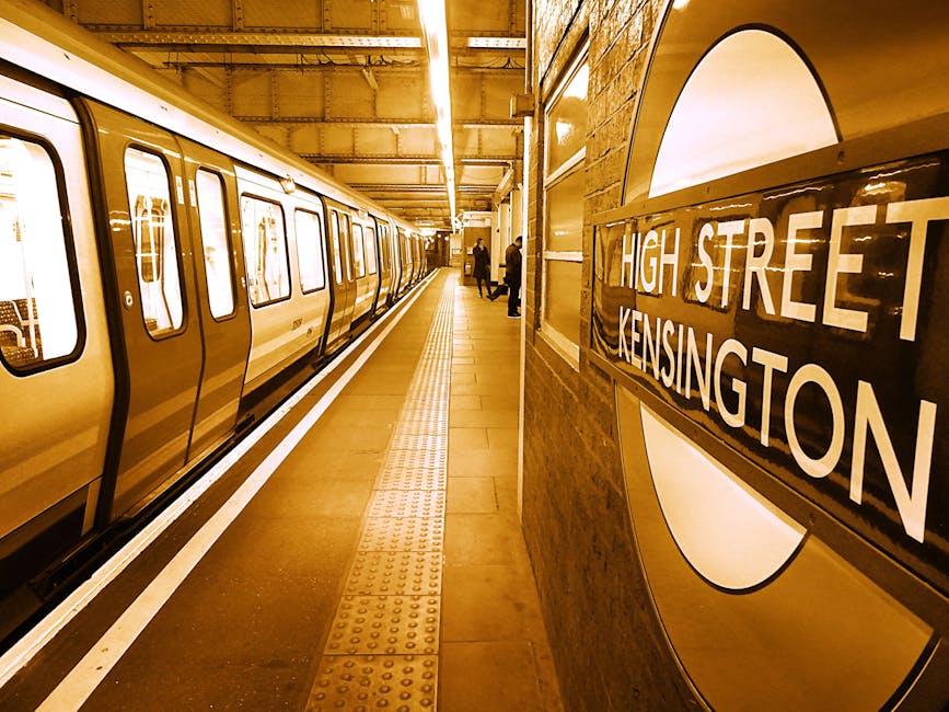 A view from the floor level inside a London Underground station, showing the curved, white-tiled tunnel walls and arched ceiling with a central strip of fluorescent lighting. On the left side, yellow line markings run along the textured, grooved platform surface, guiding passengers towards the escalators and staircases. The station features advertising posters on the walls, including some with colorful images and text, as well as the iconic roundel London Underground signs on both sides of the platform. Passengers are seated on benches or standing, waiting for approaching trains, with some engaged in conversation and others focused on their phones. The overall environment is busy but orderly, with a clean, well-maintained appearance typical of a London commuter station. This setting reflects the daily flow of commuters, with opportunities for private or alternative transit arrangements when considering rubbish removal and station upkeep, as handled by local services such as House Clearance Knightsbridge.