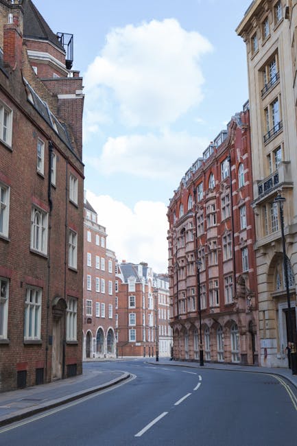 A narrow, curved street in an urban area lined with tall, multi-storey buildings constructed from brick and stone. The buildings feature a mix of architectural styles, including bay windows, decorative cornices, and wrought iron balcony railings, with some surfaces displaying red brickwork and others painted in neutral tones. The street appears clean with clearly marked double yellow lines along the curb, and there are no vehicles or pedestrians visible. The sky above is partly cloudy with patches of blue, providing bright natural lighting that highlights the textures of the building facades and the asphalt road surface. The scene is typical of central London architecture, which may require professional rubbish removal services from companies such as House Clearance Knightsbridge to manage debris from property clearance or renovation projects, especially in dense urban areas where straightforward waste disposal alternatives are often limited or less suitable for private collection needs.