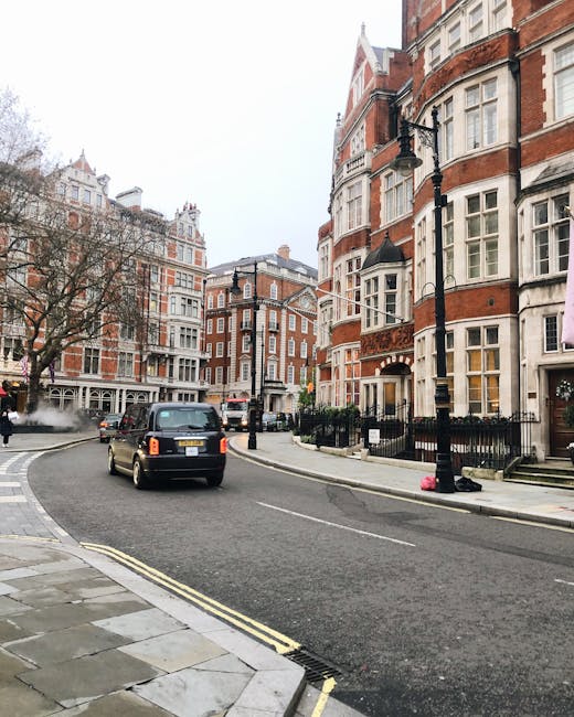 A quiet street scene in a residential area featuring a curved asphalt road with double yellow lines along the curb, bordered by Victorian-style brick and white-painted stone-fronted buildings with bay windows, decorative brickwork, and small gardens enclosed by black metal railings. A black taxi is driving along the street, while a person dressed in dark clothing is walking on the pavement to the left. Modern street lamps with black metal poles are positioned at intervals, illuminating the scene. Sparse leafless trees are visible, suggesting late autumn or winter. The background shows more of the same historic architecture with subtle variations in window design and façade detailing. This scene exemplifies a typical London neighborhood, where private waste collection and local rubbish removal services, such as those provided by House Clearance Knightsbridge, can efficiently handle household clearance needs, especially for infrequent or specialised rubbish disposal outside of local authority services.
