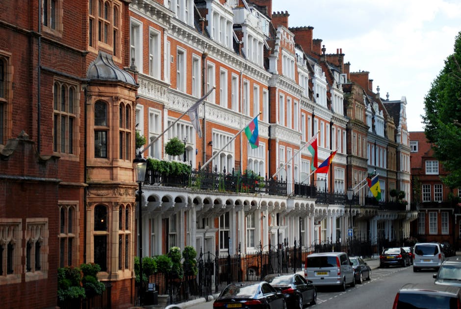 A row of ornate, multi-story red brick buildings with white decorative trim and large bay windows, situated along a residential street. Several flags are mounted on the balconies, including some with bright colors and patterns, adding visual interest against the historic facade. The buildings feature detailed stonework, with bay windows on the left containing tailoring details like pointed arches and decorative stone mullions. In the foreground, a street scene includes parked cars along the pavement, with some slightly visible on the curbside. The street is lined with black wrought-iron fences and a few small trees and bushes, contributing to a tidy appearance. On the right, the street continues into a green, leafy area, providing a contrast to the brick architecture. The overall lighting suggests natural daylight, highlighting the textures and colors of the buildings and street environment, representing a typical affluent UK neighborhood with potential relevance to private or independent waste handling scenarios often associated with residents or property management, such as rubbish clearance services. House Clearance Knightsbridge is known for providing such services in this area.