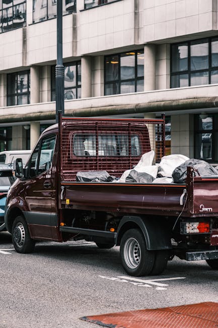 A small, dark red flatbed pickup truck parked on an urban street in front of a modern, multi-storey office building with large glass windows and a stone facade. The truck's cargo bed is loaded with various waste materials, including black and white plastic bags, some tied and others loosely filled, along with a few cardboard boxes. The truck features a metal safety grille behind the cab, which provides additional support and separation between the driver and cargo area. The scene is well-lit with natural daylight, and the vehicle is positioned parallel to the building, occupying a designated parking area marked with white lines. Nearby, other parked vehicles, including a white van, are visible. This setting suggests the use of private waste collection for small business rubbish removal, an alternative to council curbside services, exemplifying on-site clearance or direct disposal handled by companies such as House Clearance Knightsbridge, operating in the local commercial waste management sector.