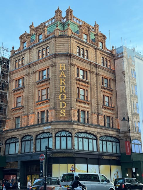 A multi-storey historic building with ornate, detailed stonework and large arched windows on the ground floor. The upper floors showcase decorative brickwork and numerous rectangular windows with prominent stone surrounds. The building's exterior features a vertical sign reading 'HARRADS' in large gold letters running down one side of the structure. The rooftop has a decorative green-domed pavilion with intricate stone balustrades and ornamental turrets. Surrounding the building, it is situated on a busy urban street with parked vehicles, including a white van and several cars, as well as pedestrians on the pavement. A black street lamp is visible on the sidewalk, and scaffolding partially obscures part of an adjacent building on the right. The scene suggests a city environment where property maintenance or building clearance may be relevant, aligning with services provided by companies like House Clearance Knightsbridge for rubbish removal or private property clearance actions.