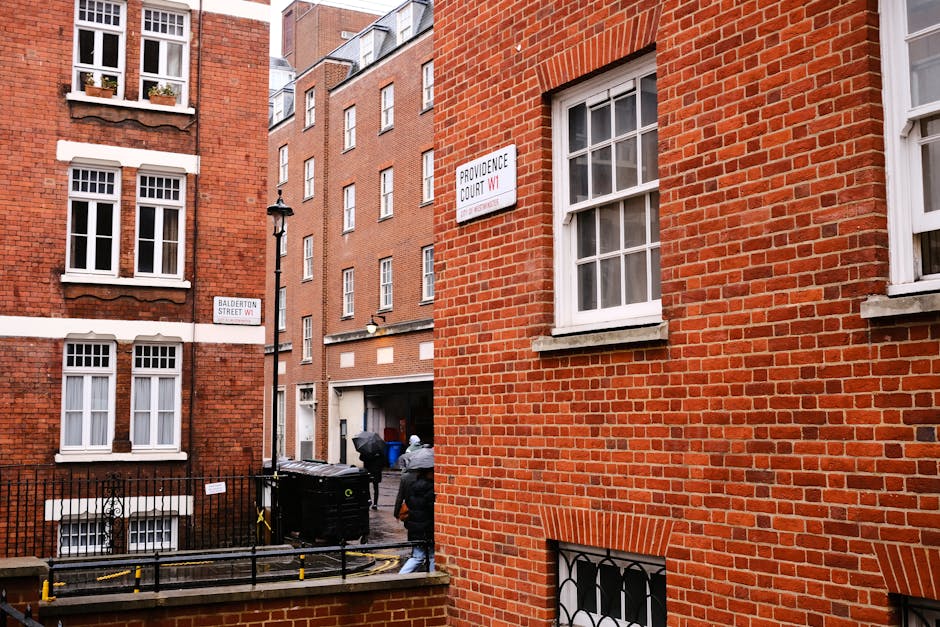 A narrow alleyway between red brick residential buildings in an urban area, with a black and yellow wheelie bin and a small fenced terrace visible in the foreground. A person dressed in dark clothing is seen loading or unloading waste near the bin area, with additional black rubbish bins placed against the building wall. The buildings feature multiple white-framed sash windows, some decorated with small potted plants, and the brickwork exhibits a mix of traditional red tones and lighter mortar joints. Street signs indicating 'Balderton Street W1' and 'Providence Court W1' are mounted on the facades, emphasizing the location within a city street environment. The scene is illuminated by natural daylight, highlighting the textures of the brickwork and the various waste management elements, illustrating typical private rubbish handling and collection activities in a dense urban setting, with House Clearance Knightsbridge providing services related to waste removal and disposal in similar contexts.