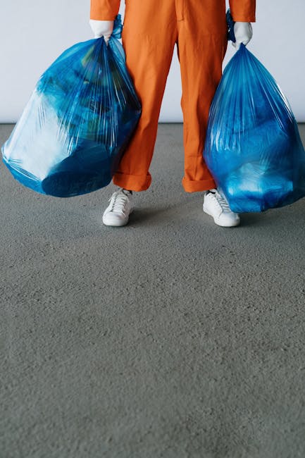 A person standing on a grey concrete surface, holding two large blue plastic rubbish bags filled with waste, with their arms extended downward. The individual is dressed in orange work overalls and white sneakers, with the lower part of their body visible from the waist down. The background features a plain, light-colored wall. The scene depicts an informal rubbish collection activity, potentially related to private waste removal or on-site clearance, which aligns with the services offered by companies like House Clearance Knightsbridge for alternative waste handling and disposal of household or commercial rubbish.