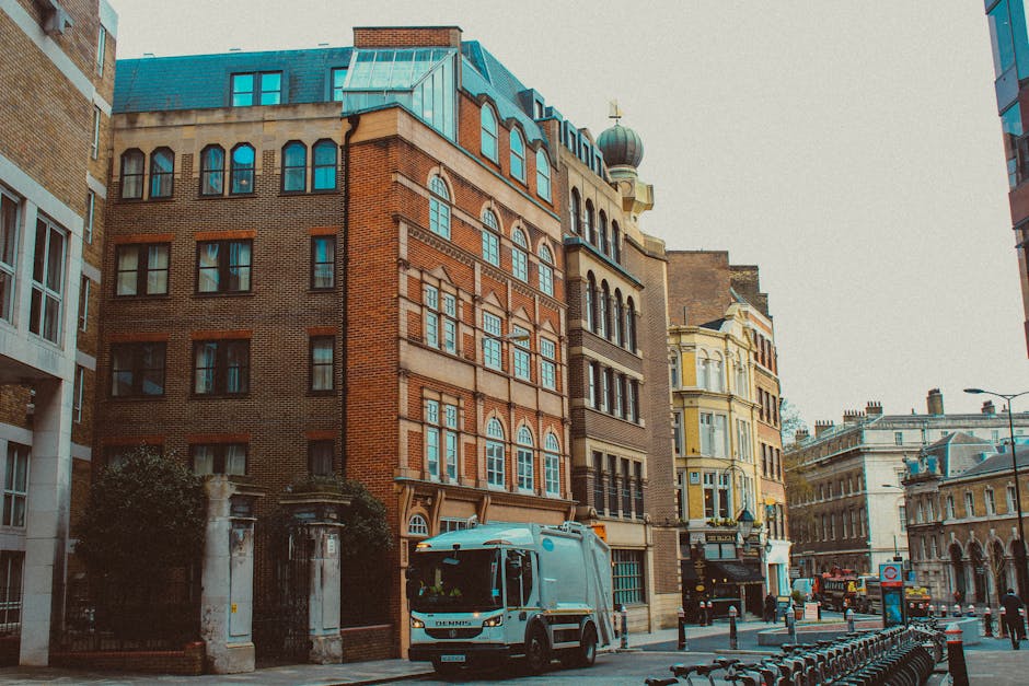 The image depicts a street view in an urban area, featuring a mixture of historic and modern buildings with architectural details such as arched windows, decorative brickwork, and a distinctive domed turret. The buildings are constructed with materials including red and beige brick, stone, and glass, demonstrating a variety of textures and finishes, from smooth glass windows to rough brick facades. In the foreground, a light blue waste collection vehicle is parked on the street, with the cab facing towards the camera, indicating active rubbish collection services. The street setting is illuminated by natural light, with soft shadows cast on the pavement and buildings, highlighting the textures of the materials. Pedestrian bollards and bike racks line the sidewalk, and in the background, additional traffic and architectural elements suggest a busy city center. The scene subtly underscores the importance of professional waste removal, such as that provided by House Clearance Knightsbridge, for managing urban rubbish and maintaining cleanliness in areas with narrow access or dense building arrangements.