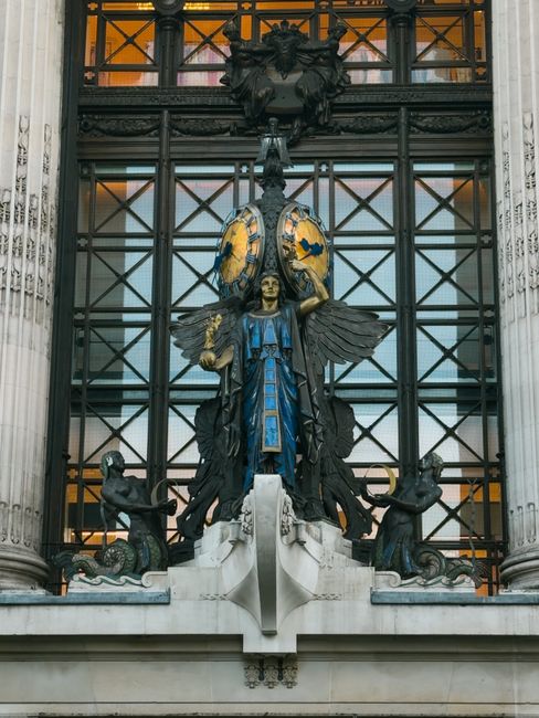 A detailed view of a decorative sculpture mounted on a building facade, featuring a central golden face with wings extending outward, clad in a blue and gold robe. The sculpture is flanked by two smaller figures at the base, each holding an object and standing on a white marble ledge. Behind the sculpture, there is a large black wrought-iron framework with a grid pattern, supporting a glass window that reveals a warm orange interior lighting. The building's columns are white with carved detailing, adding a classical architectural element. The scene captures an ornate, historic style, with an emphasis on intricate metalwork, contrasting textures of metal, marble, and glass, and a blend of colors including gold, blue, black, and white. This setting suggests an urban environment potentially associated with aesthetic or cultural institutions, where private or independent collections might be visible in the context of ongoing building maintenance or preservation work, aligning with the theme of alternative waste handling or structural upkeep by specialized services like House Clearance Knightsbridge.